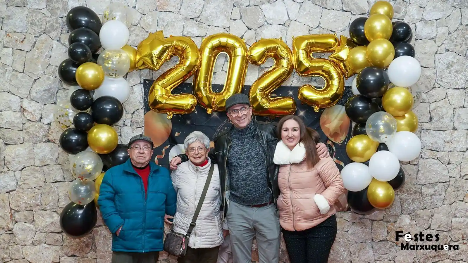 Grupo de adultos mayores y una joven posan sonrientes junto a los globos dorados 2025 en la celebración de Nochevieja de Marxuquera