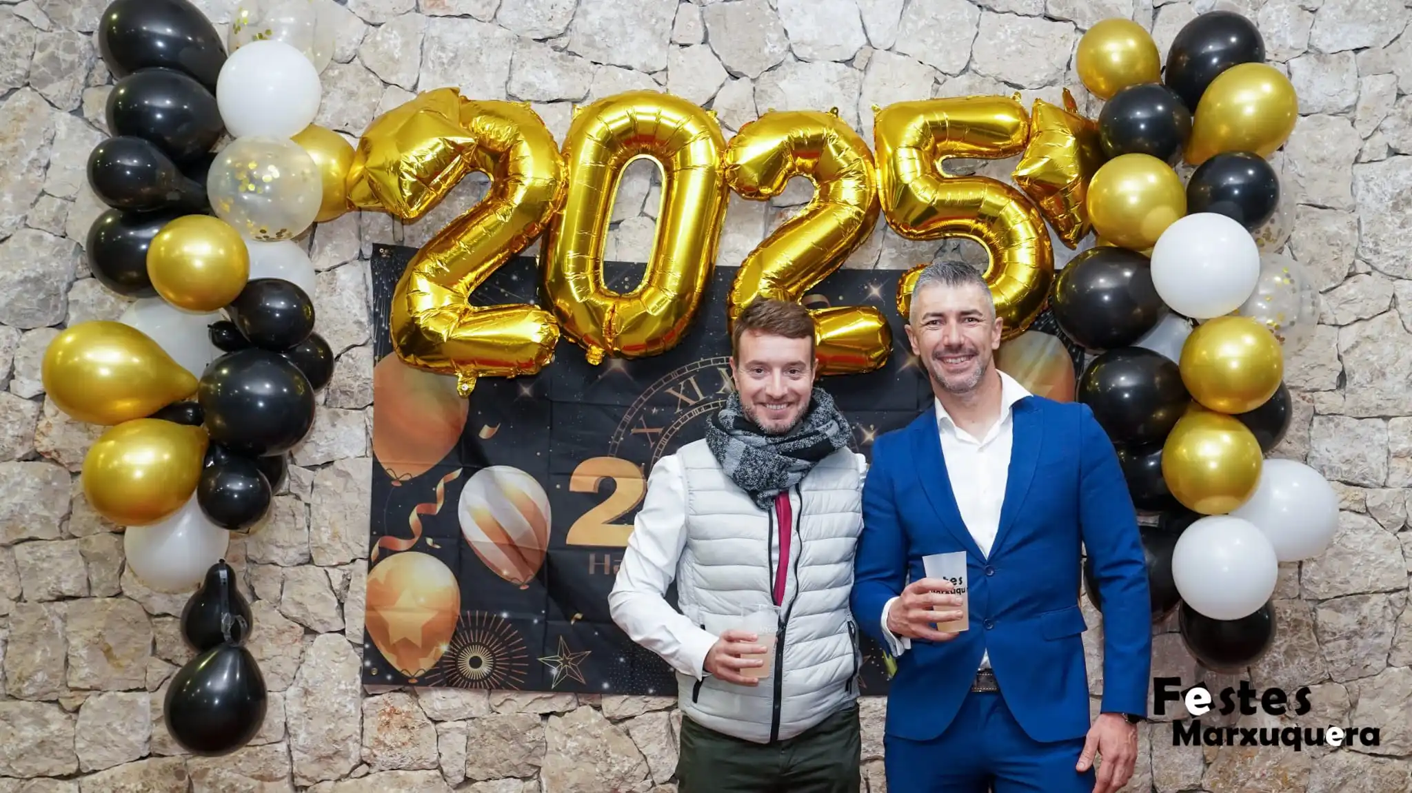 Dos hombres posan sonrientes con copas en la mano junto a los globos de año nuevo en Marxuquera