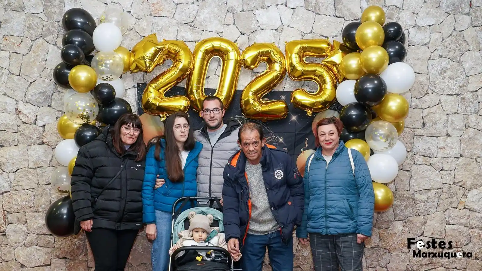 Grupo familiar con bebé en carrito posa frente al mural decorado con globos dorados de 2025 en el Centre Social de Marxuquera