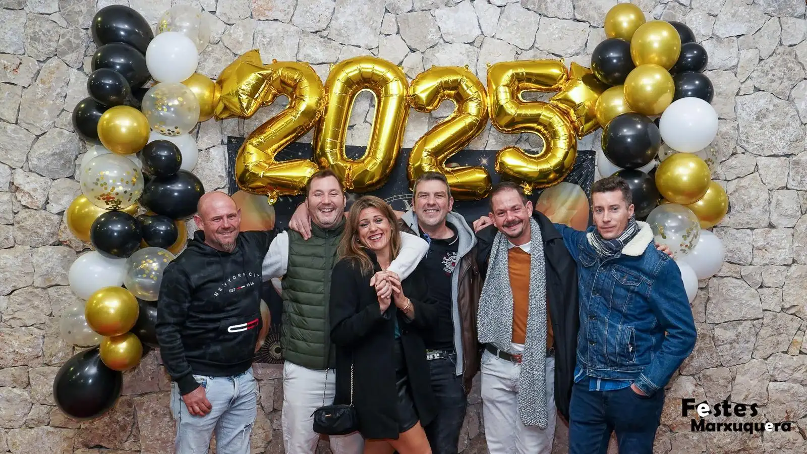 Grupo de seis amigos sonríe bajo los globos dorados de 2025 en la fiesta de Nochevieja del Centre Social de Marxuquera