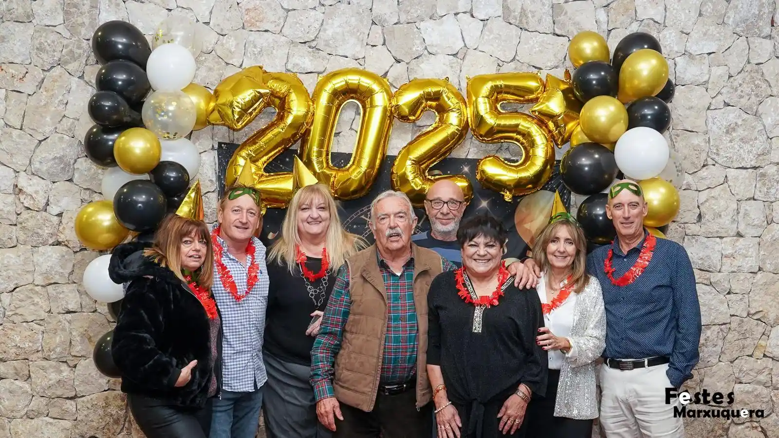 Grupo de ocho personas con gorros y collares festivos posa junto a la decoración de globos dorados en la Nochevieja de Marxuquera