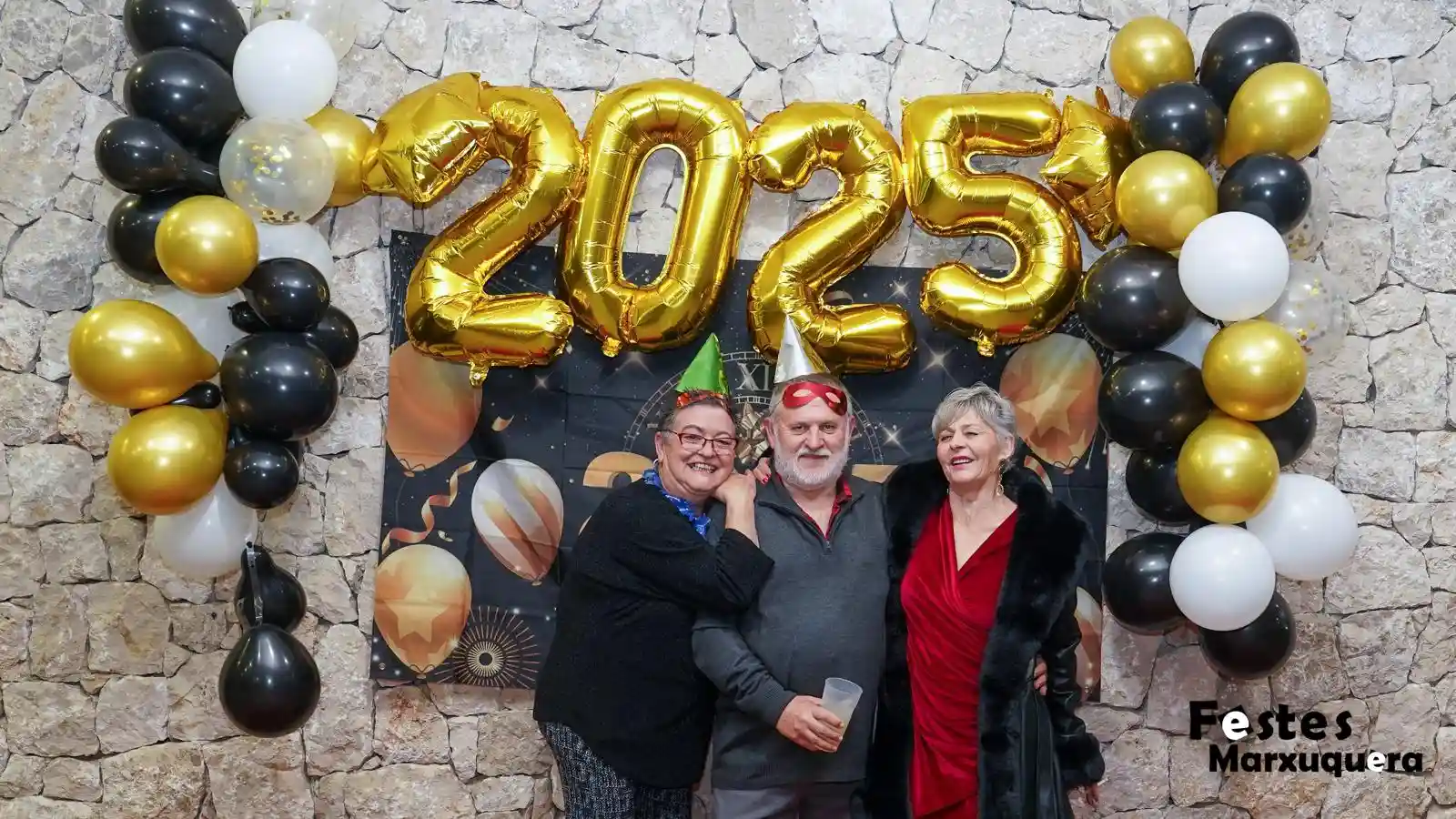 Un hombre con gafas y dos mujeres con gorros de fiesta posan ante el decorado 2025 de la Nochevieja en Marxuquera