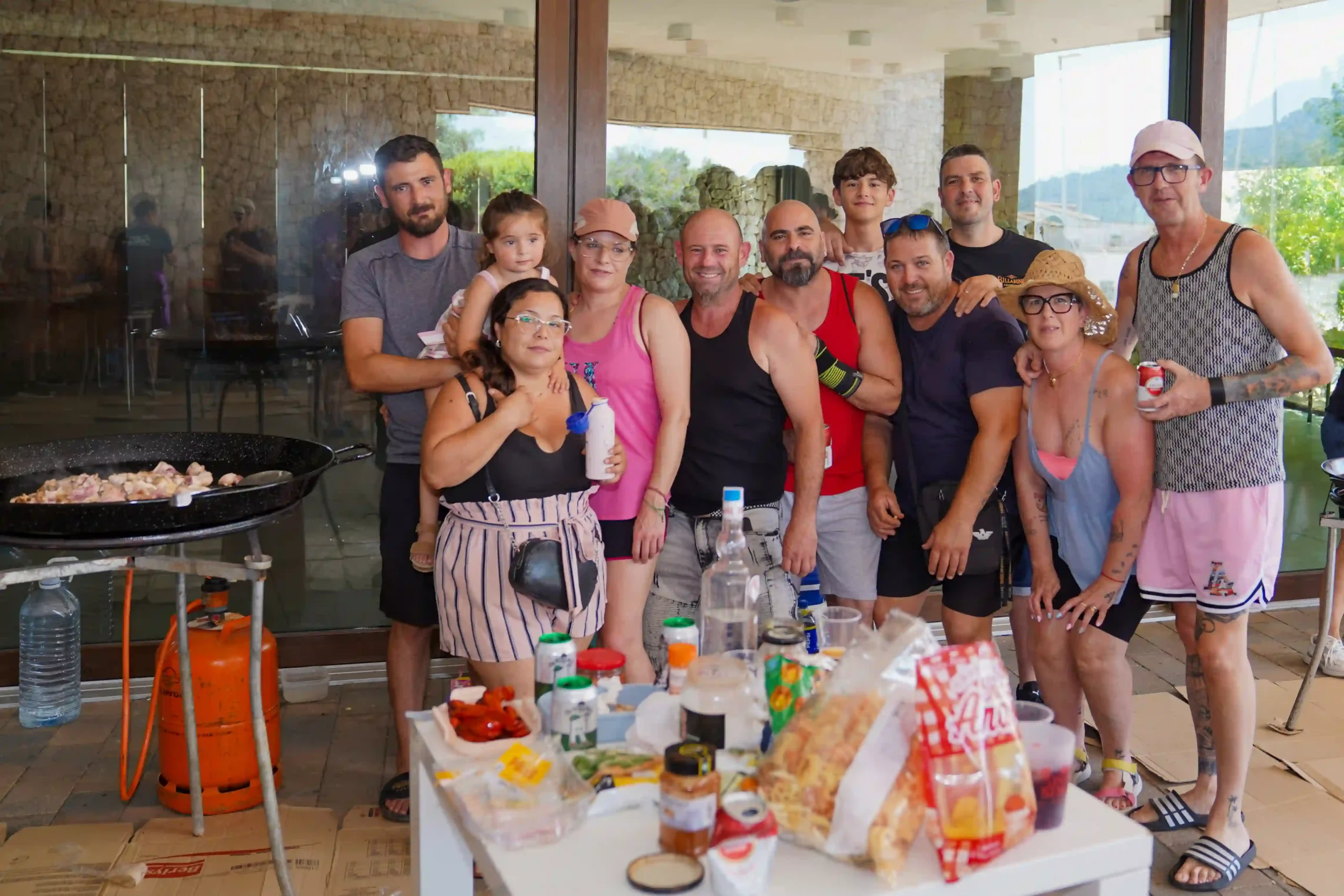 Grupo numeroso disfrutando de comida y cocinando paella con carnes durante la Romería a la Mare de Déu del Mondúver 2025 en Marxuquera, junto a cristalera y pared de piedra