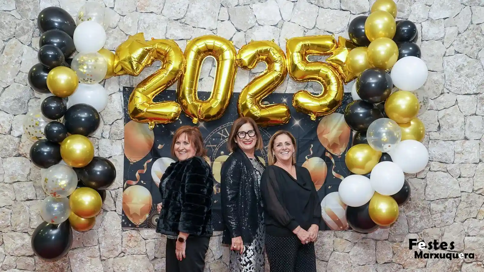 Tres mujeres vestidas de gala posan sonrientes junto a la decoración de globos y números 2025 en el photocall de Nochevieja