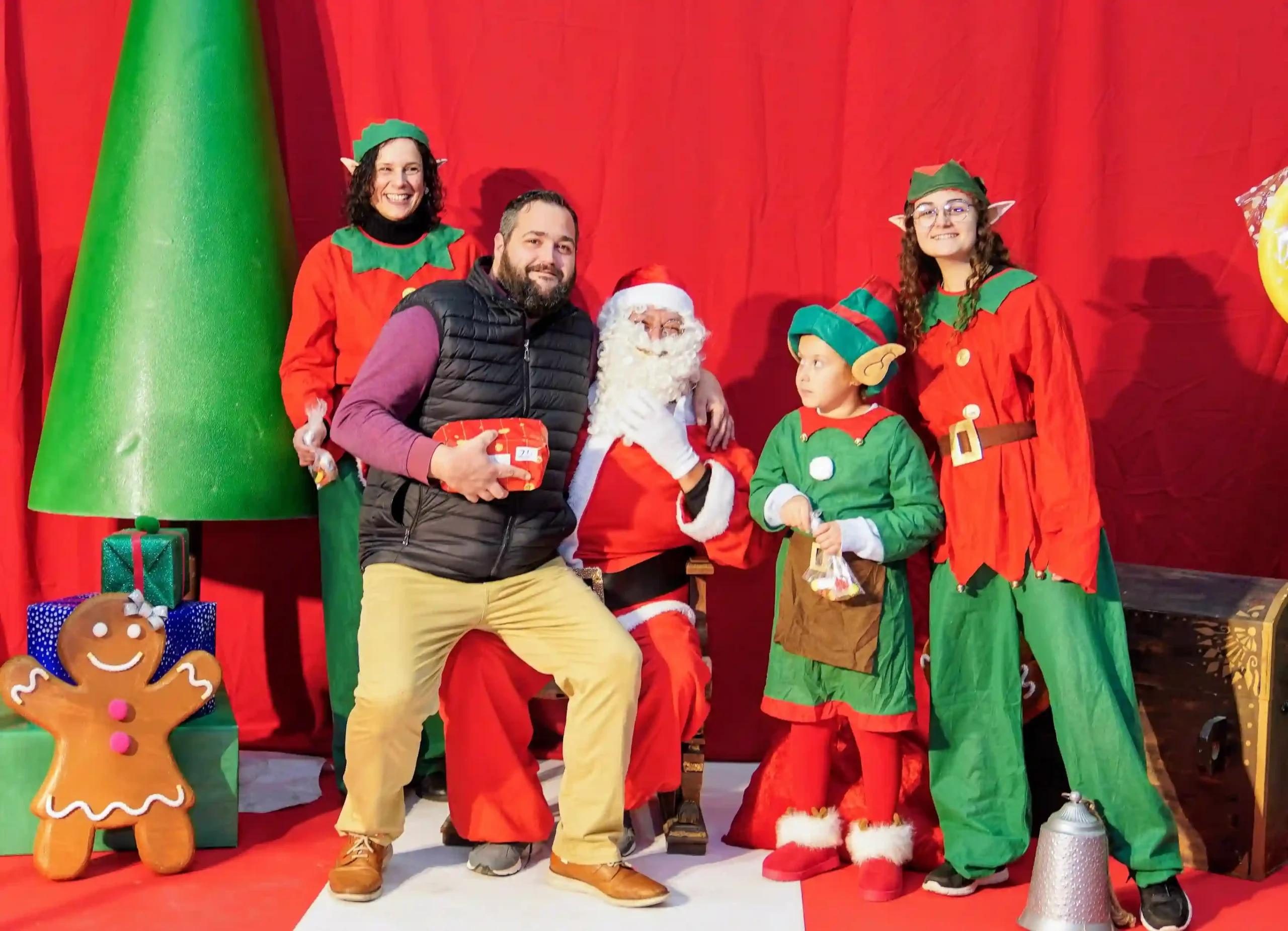 Hombre recibiendo un regalo y posando con Papá Noel y sus elfos en el decorado navideño