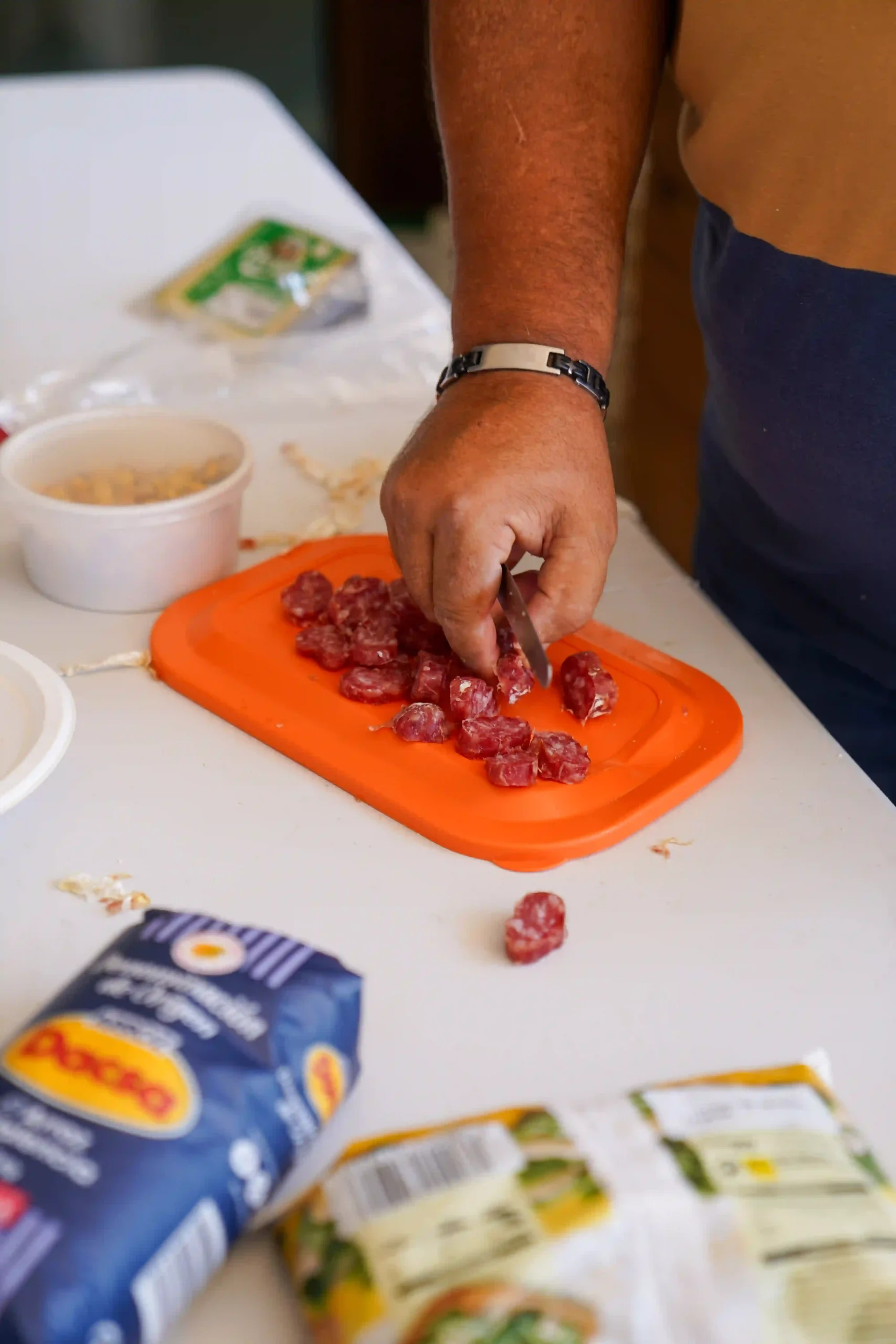 Persona cortando fuet sobre tabla naranja junto a ingredientes tradicionales como arroz Dacsa, garbanzos y verduras, durante la Romería a la Mare de Déu del Mondúver 2025 en Marxuquera